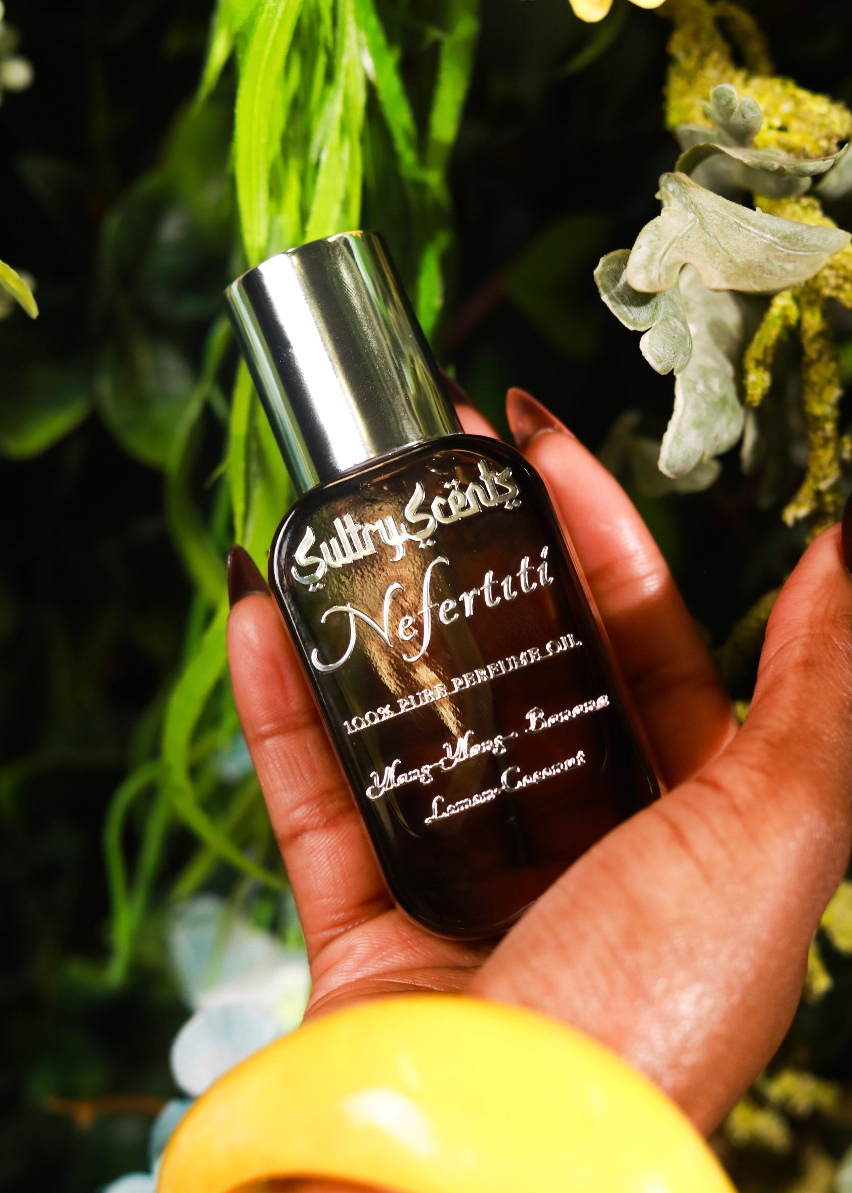 Hand holding SultryScent Nefertiti perfume oil bottle amidst lush greenery, representing luxury and lasting fragrance.